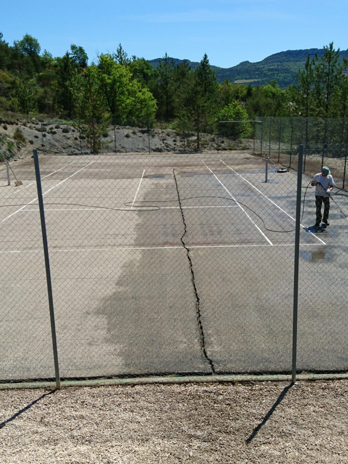Court de tennis avec une personne en train de jouer, entouré d'une clôture et d'arbres, avec des montagnes en arrière-plan.