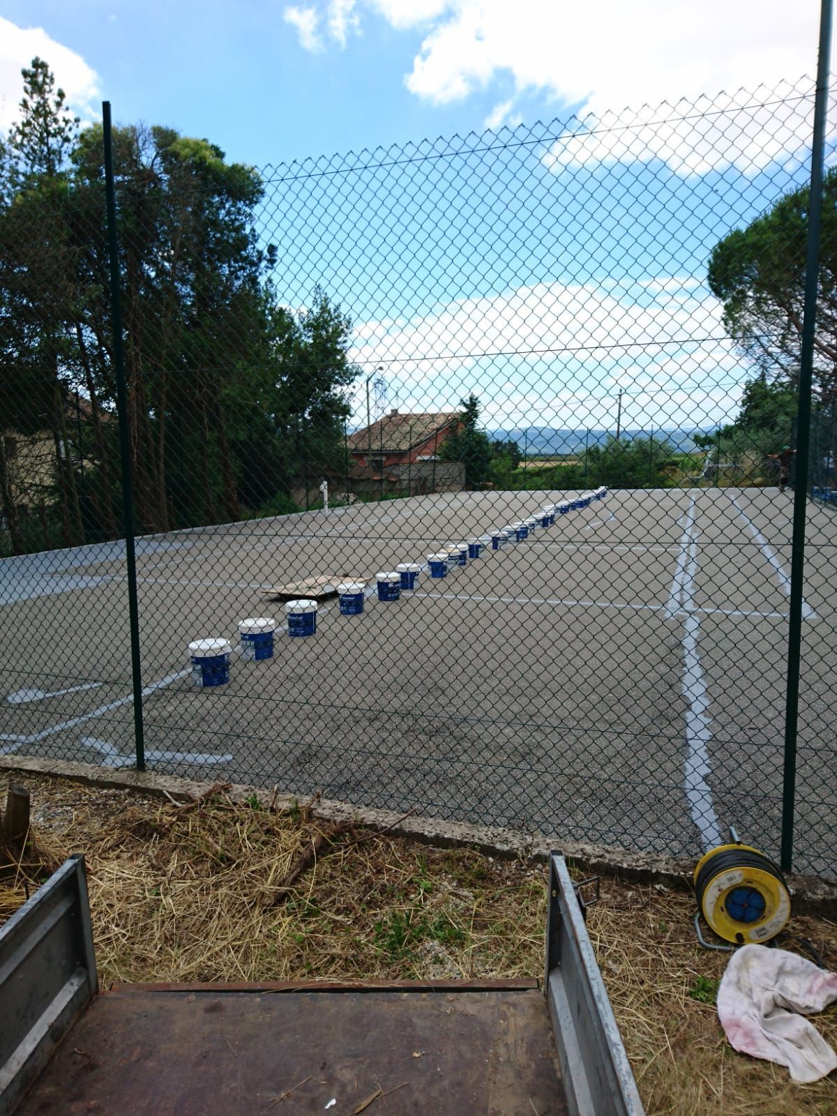 Un court de tennis est en cours de peinture ; on aperçoit, à travers une clôture, des seaux de peinture bleue alignés.