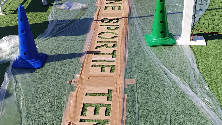 Des cônes bleus et verts encadrent un panneau en carton sur lequel on peut lire TENNIS SPORT ONE, le tout sur une pelouse verte recouverte de plastique.
