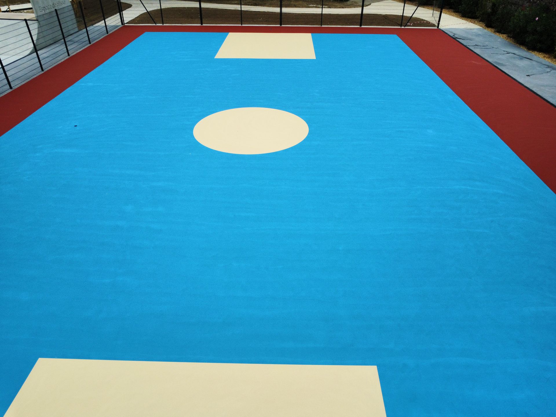 Terrain de sport bleu et beige à bordure rouge, espace extérieur clôturé.