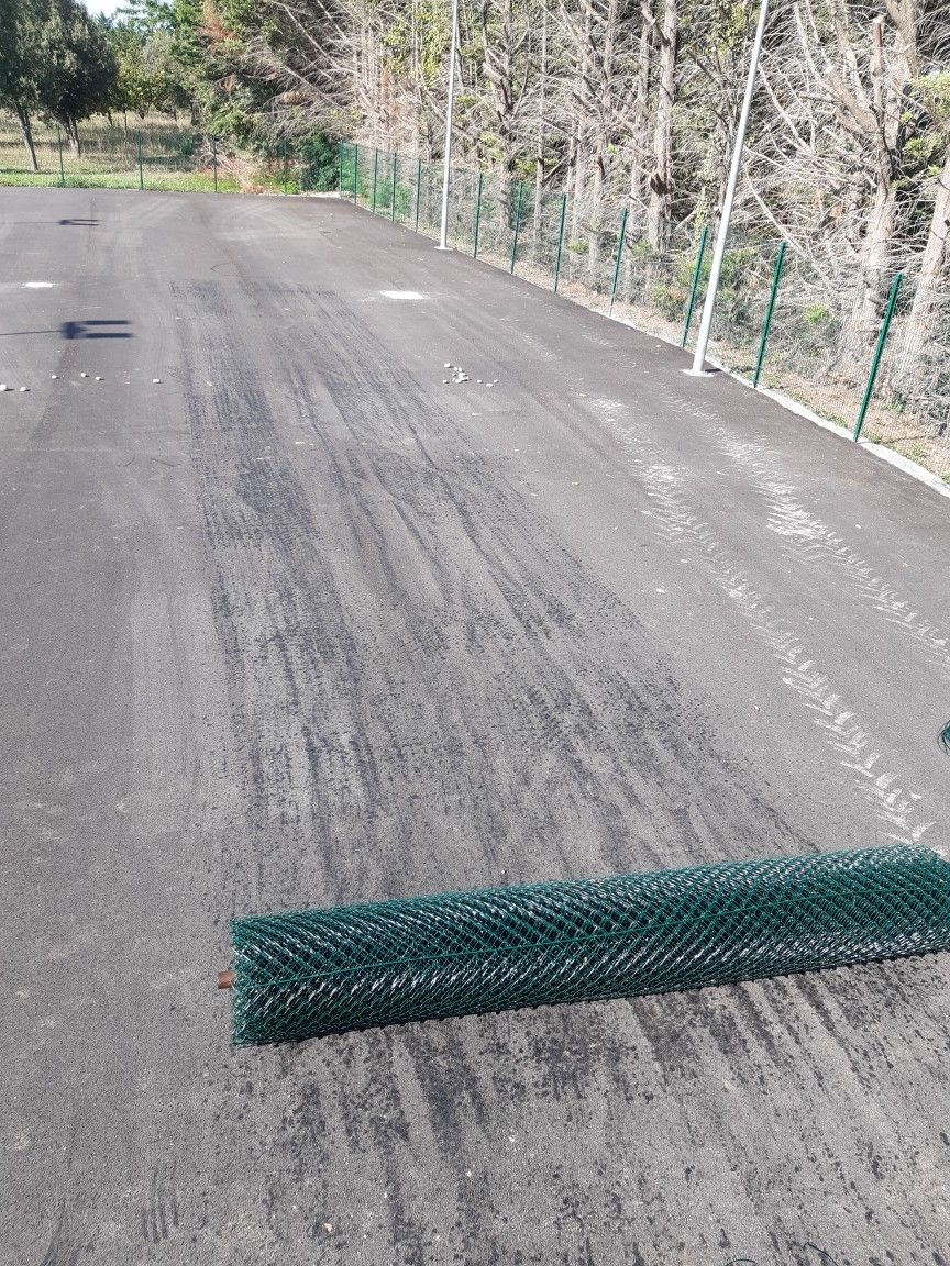 Route en asphalte foncé bordée d'une clôture verte ; arbres et clôture en arrière-plan.