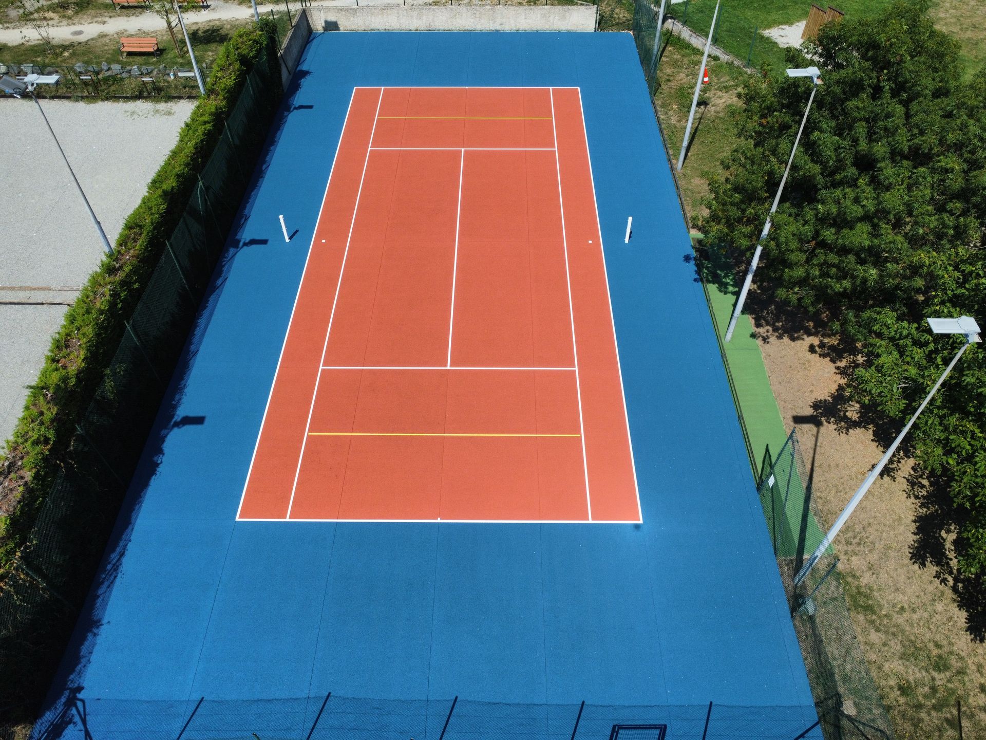 Vue aérienne d'un court de tennis bleu et rouge aux lignes blanches, entouré de verdure et de lumières.