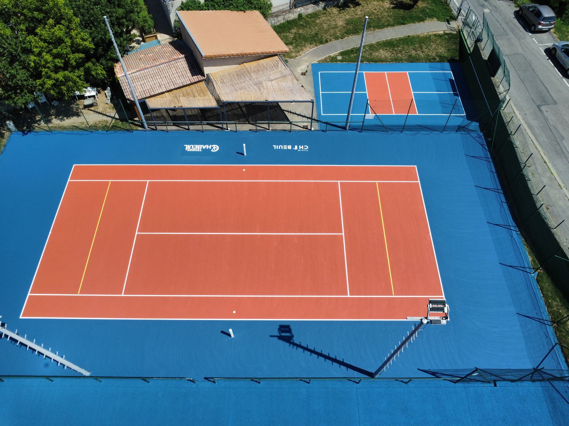 Vue aérienne d'un court de tennis bleu et orange situé à côté d'un autre court plus petit, également bleu et orange, près de bâtiments et d'une route.