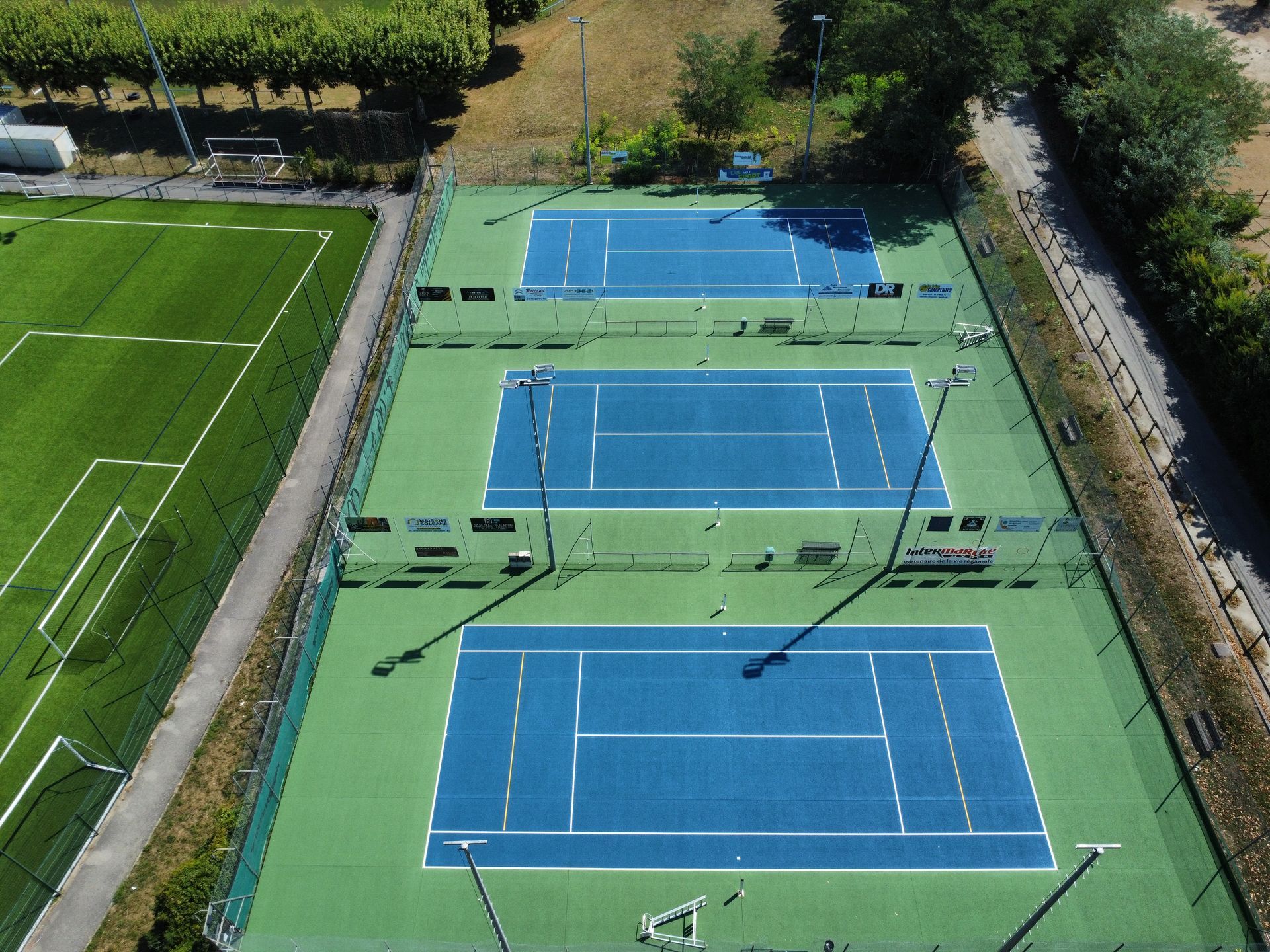 Vue aérienne de trois courts de tennis bleus, d'un champ vert et d'un chemin sous un soleil radieux.