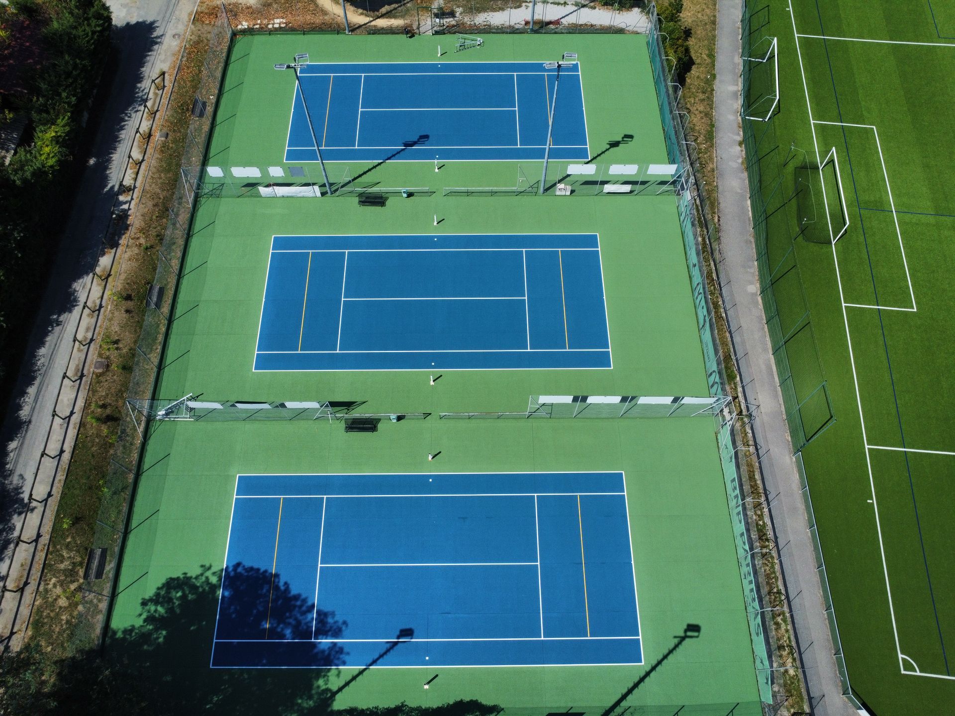 Vue aérienne de trois courts de tennis bleus, d'un environnement verdoyant et d'un terrain de sport.