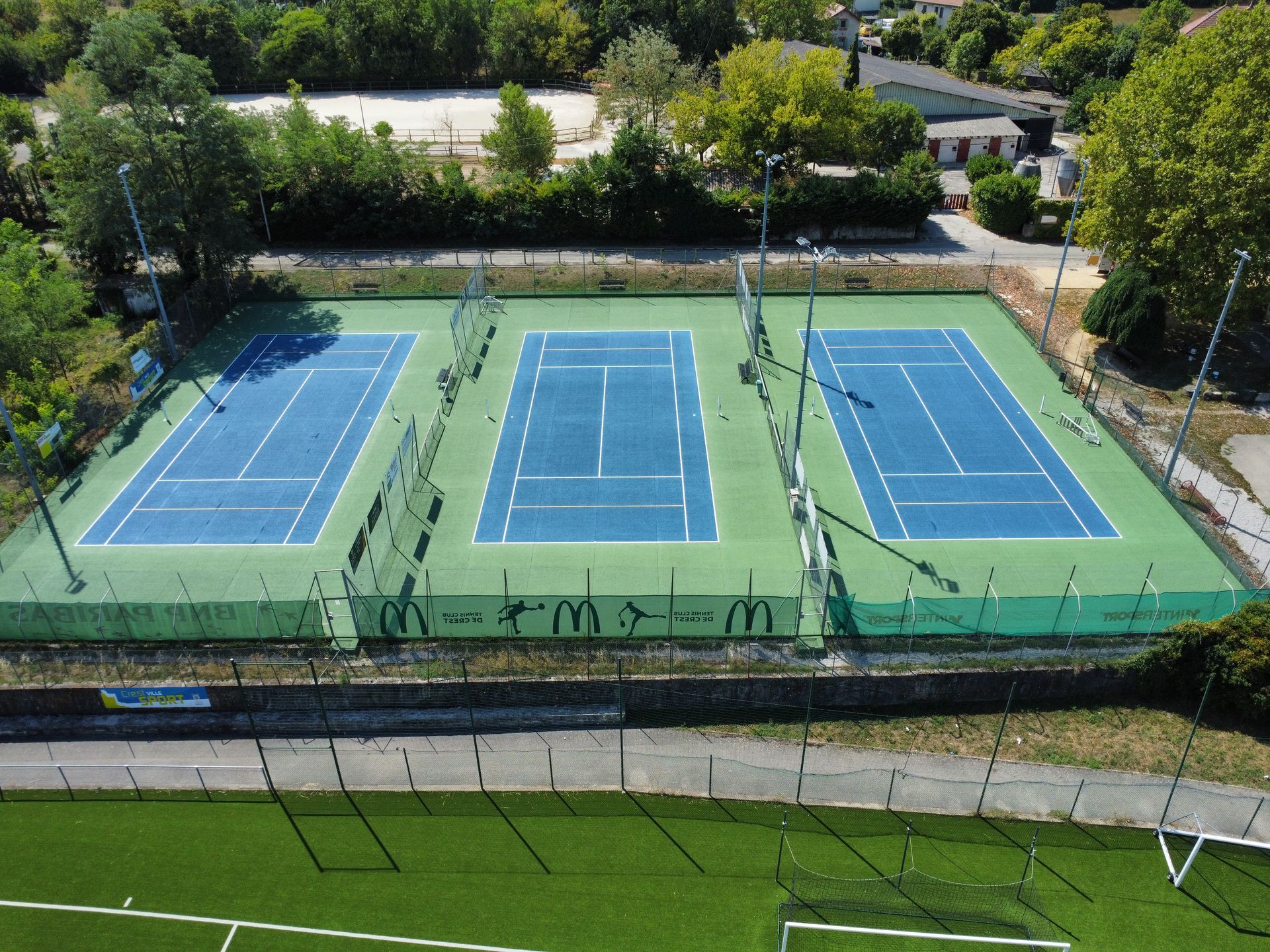 Trois courts de tennis bleus à lignes blanches sur surface verte, entourés d'arbres et d'une clôture.
