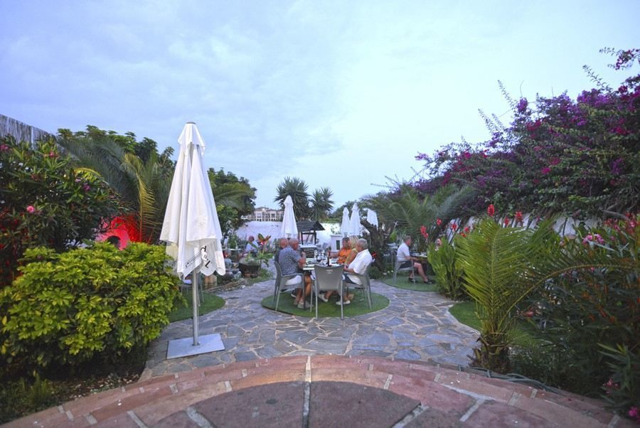 Patio exterior de piedra con sombrillas blancas, mesas y exuberantes arbustos verdes al atardecer.