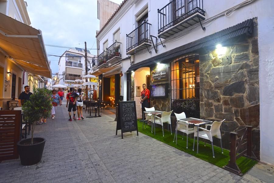 Calle estrecha empedrada con cafés al aire libre y edificios blancos al atardecer.