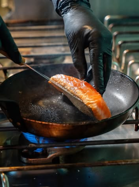 Ein Koch wendet in der Pfanne einen Lachs und brät diesen an