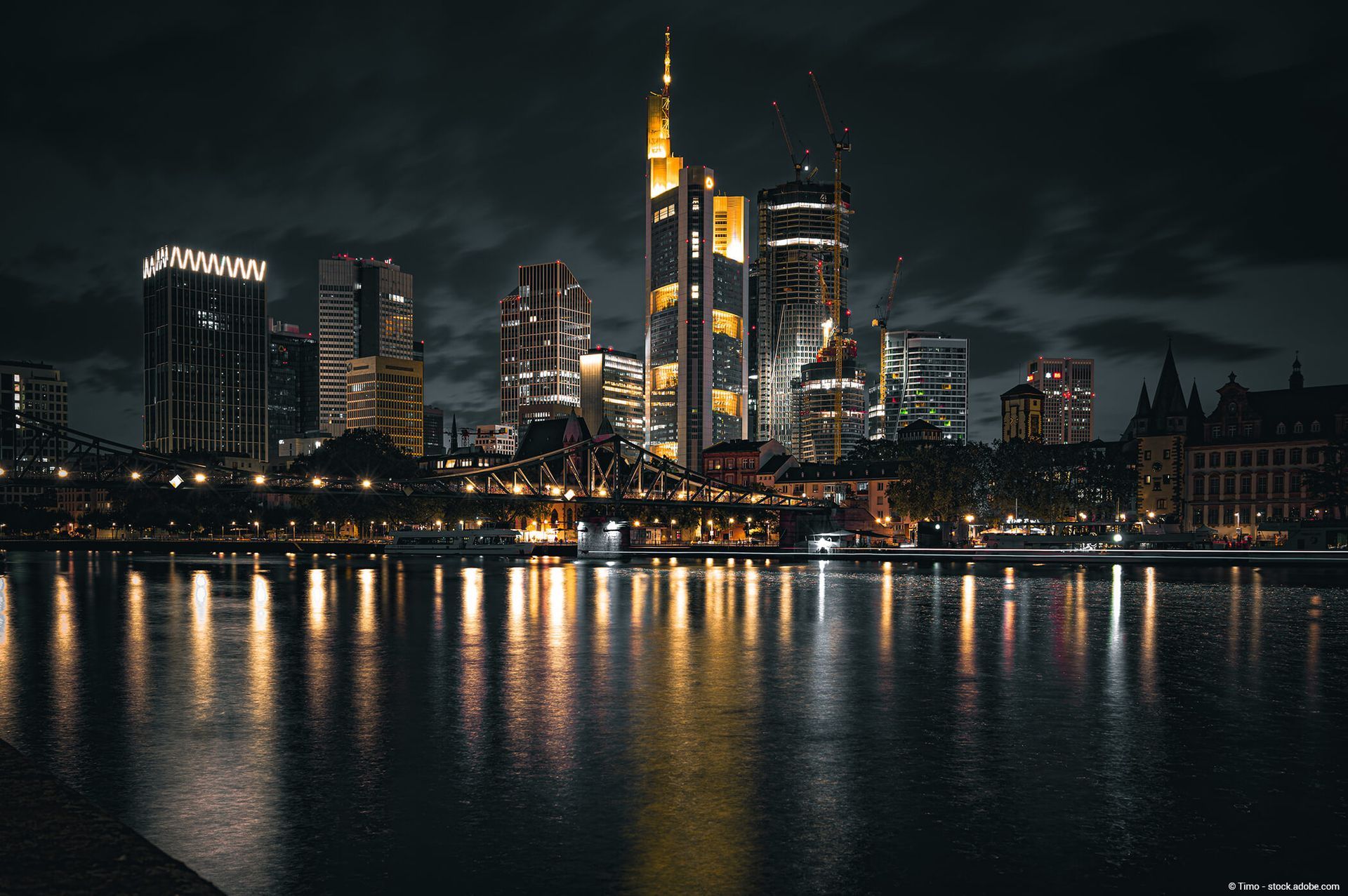 Nachtansicht der Frankfurter Skyline, die sich in einem dunklen Fluss spiegelt, die Gebäude sind gelb und weiß beleuchtet.