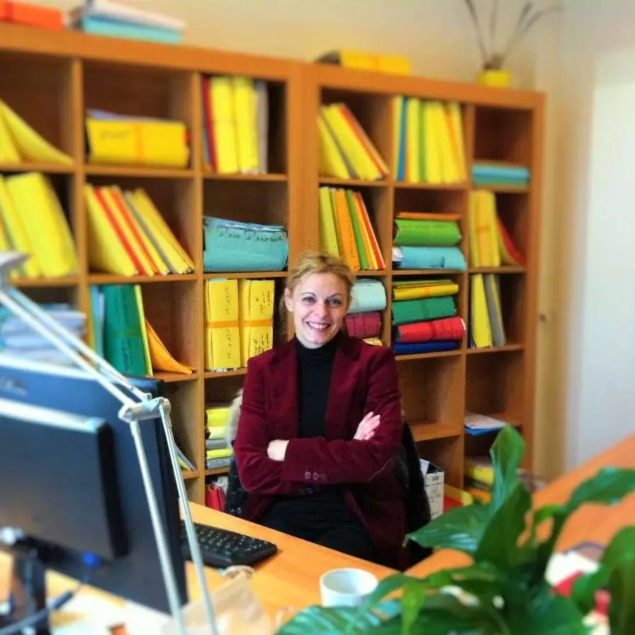 Une femme en blazer rouge sourit à son bureau, devant des étagères remplies de dossiers.