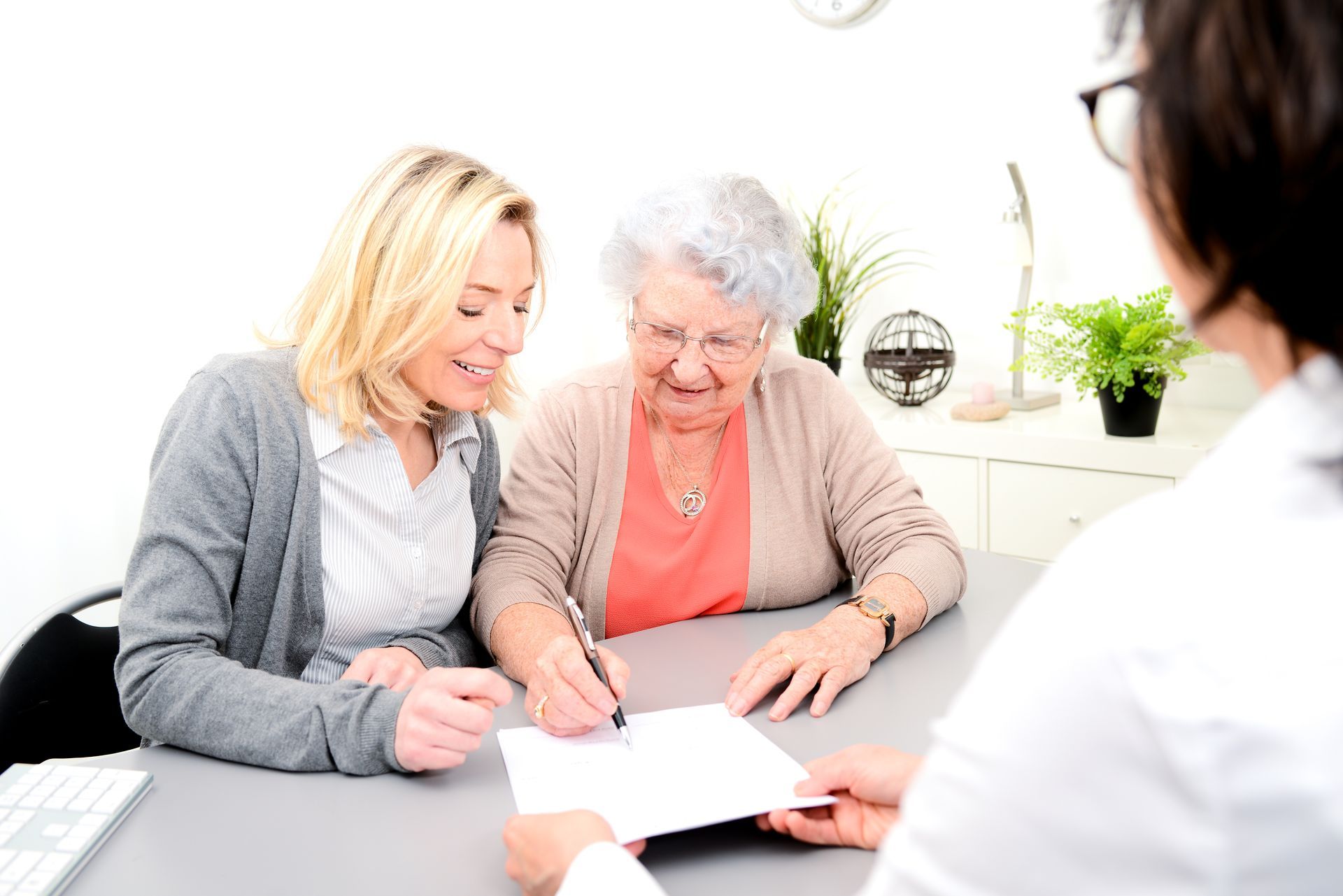 Une femme signe un document assistée d'une autre femme ; elle est assise à une table avec une personne en blouse blanche.