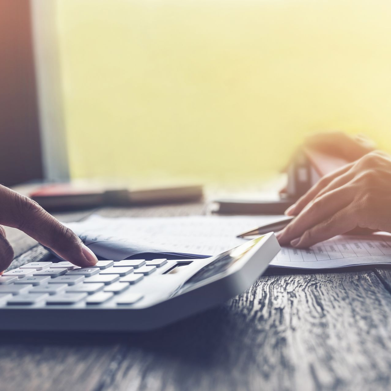 Des mains manipulent une calculatrice et un stylo sur des documents posés sur une table en bois. La lumière du soleil brille.