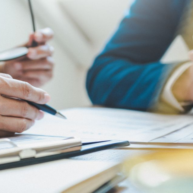 Des personnes examinent des documents autour d'une table. L'une tient des lunettes, l'autre écrit au stylo.