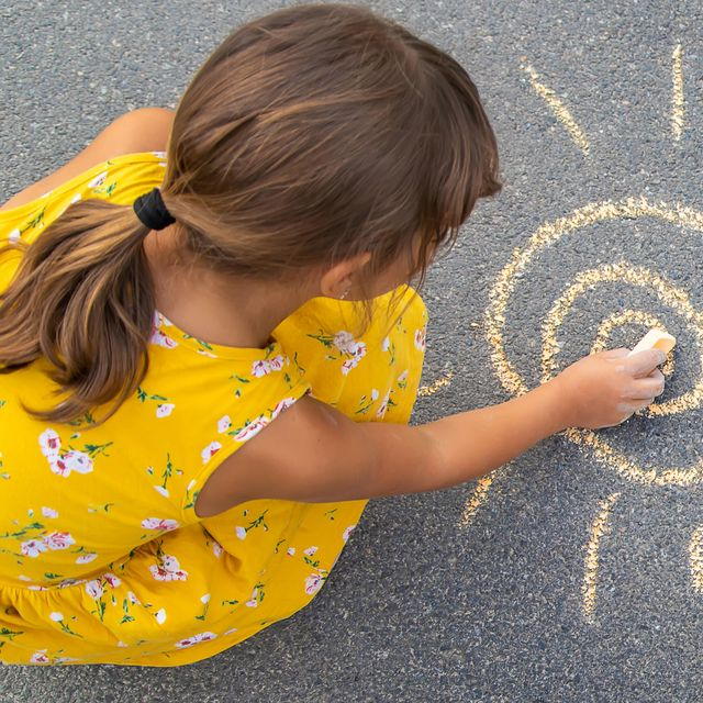 Une jeune fille en robe jaune dessine un soleil à la craie sur le trottoir.
