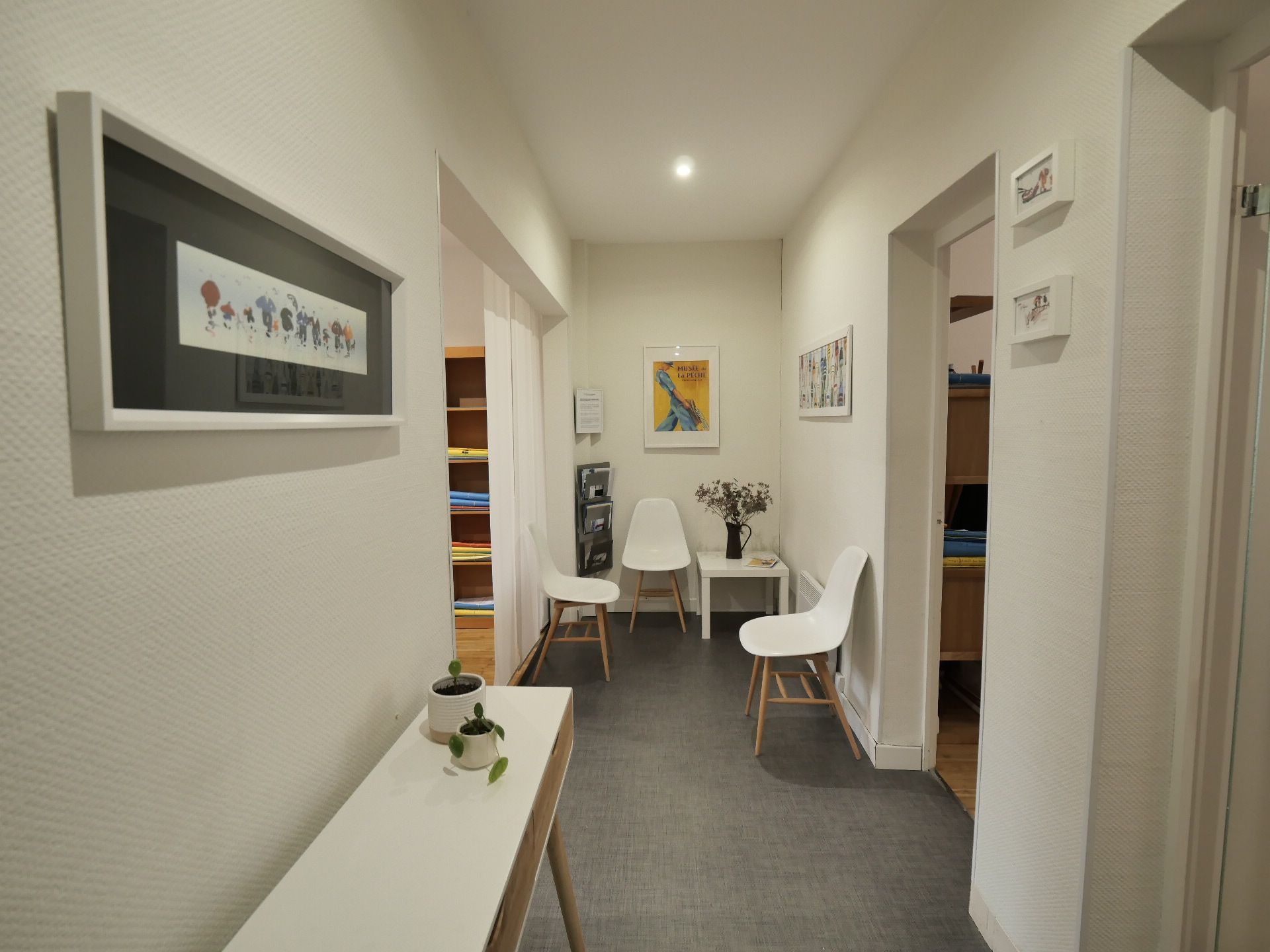 Couloir étroit aux murs blancs, tableaux encadrés, chaises d'attente et console avec plantes.