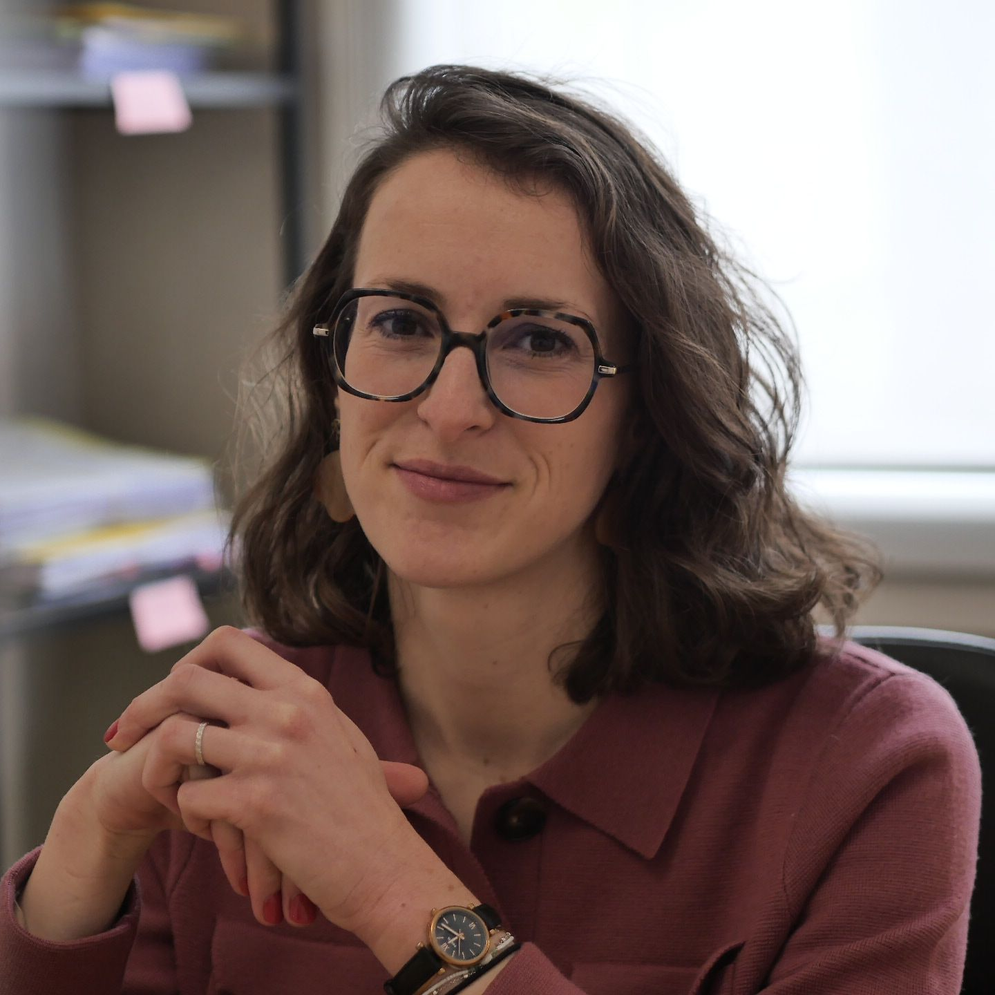 Femme aux cheveux bruns ondulés, portant des créoles, du rouge à lèvres foncé et un blazer noir, souriante.