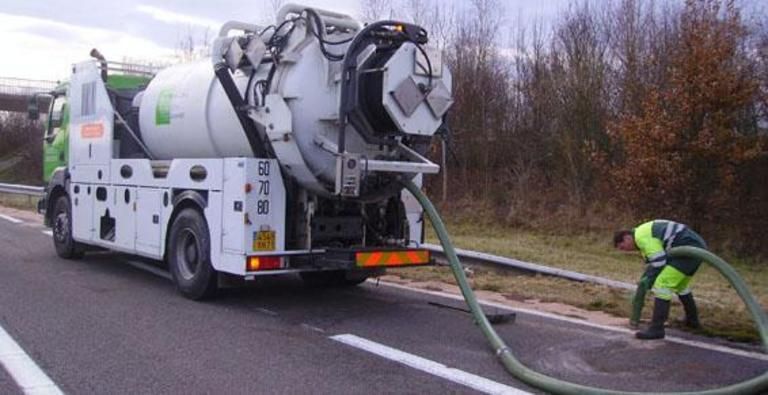 Un camion-benne et un employé aspirent des débris en bord de route. Le camion est blanc et vert.