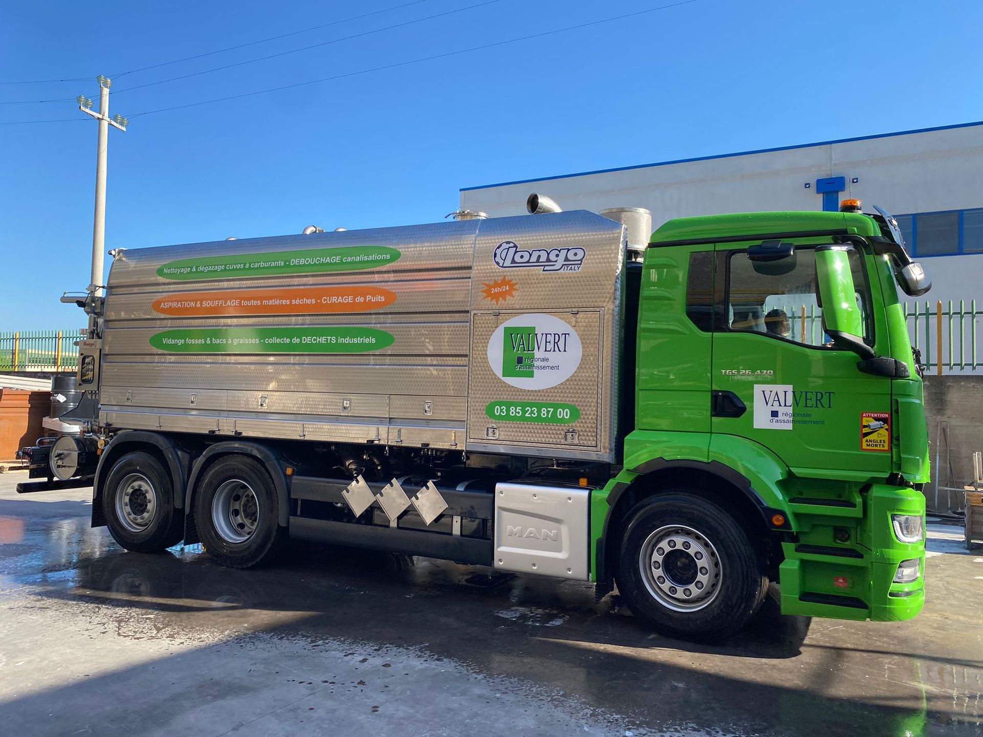 Camion de ramassage des ordures vert et blanc stationné dans une rue devant des immeubles, tuyau d'arrosage déployé.