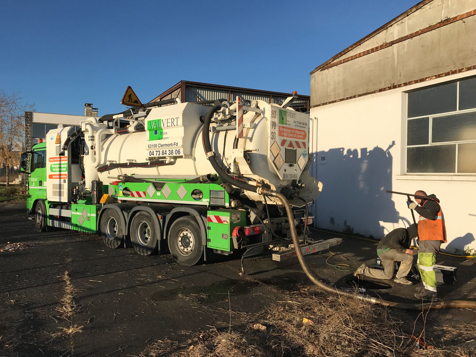 Camion-poubelle vert et blanc stationné devant un bâtiment gris par une journée ensoleillée.