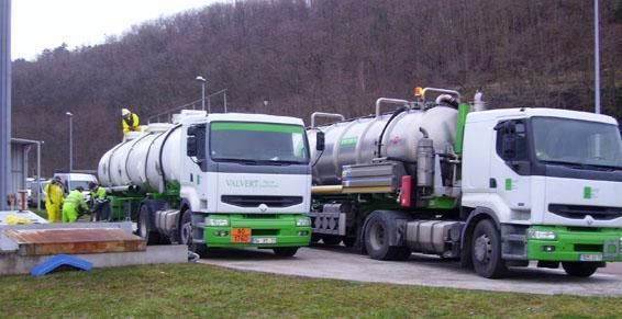 Deux camions-citernes à bordures vertes sont stationnés sur le site d'une installation ; on aperçoit des ouvriers en tenue de protection jaune.