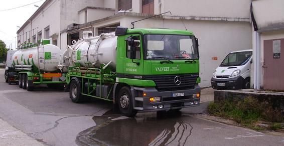 Un camion-citerne vert avec une remorque, stationné près d'une camionnette blanche et d'un bâtiment.