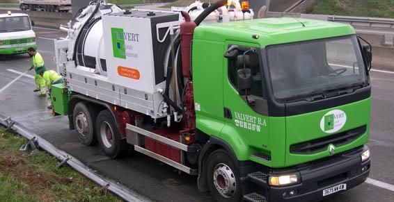 Un camion aspirateur vert et blanc nettoie une route, avec des ouvriers en gilets jaunes.