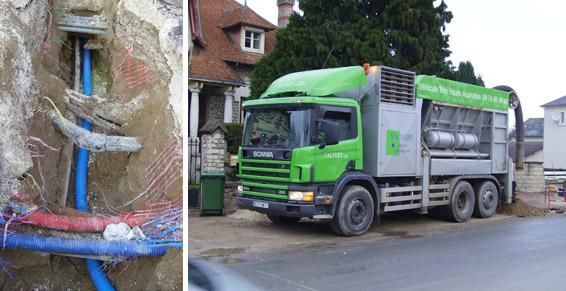 Tranchée avec des tuyaux et des câbles bleus à côté d'un camion utilitaire vert stationné dans une rue près d'une maison.