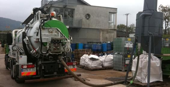 Un camion-citerne déverse des déchets dans un grand sac sur le site d'une installation, près de barils et d'un bâtiment.