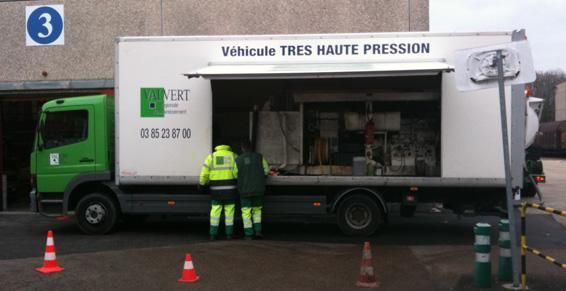 Camion vert portant l'inscription « Véhicule TRÈS HAUTE PRESSION », deux ouvriers en gilets haute visibilité, chargement/déchargement. Extérieur, sur un sol en béton.