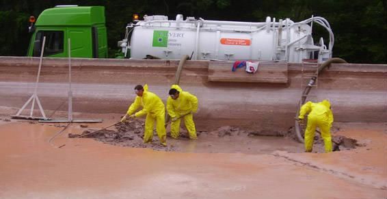 Trois ouvriers en combinaisons jaunes nettoient une fosse boueuse à l'aide de pelles, un gros camion se dresse en arrière-plan.