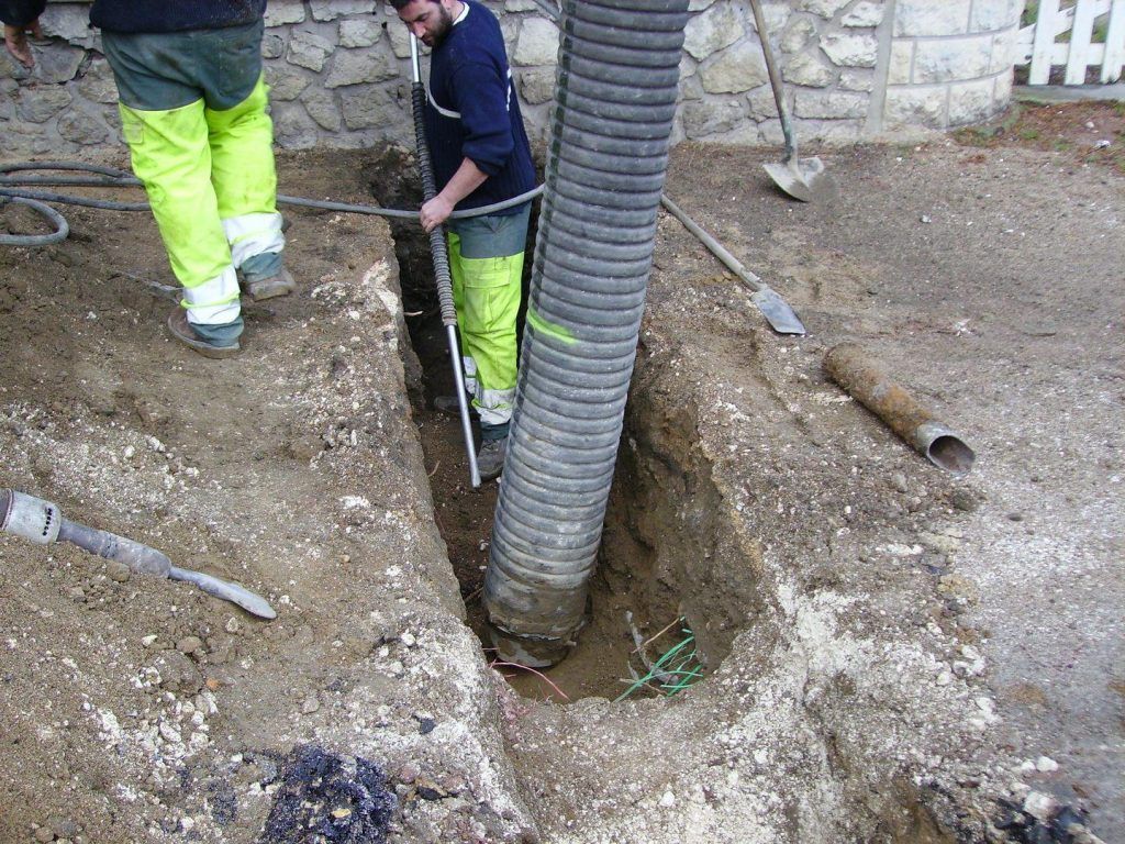 Deux ouvriers installent un gros tuyau dans une tranchée à l'extérieur. L'un tient le tuyau, l'autre se tient à proximité.