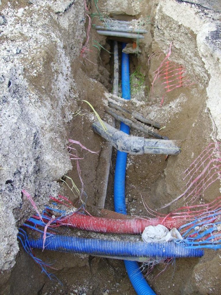 Des conduits et des câbles bleus dans une tranchée excavée, avec un filet de protection rouge et rose et une structure en béton gris.