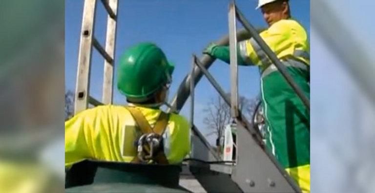 Deux ouvriers portant des gilets de sécurité et des casques de chantier travaillent sur une structure métallique, l'un d'eux grimpant à une échelle.