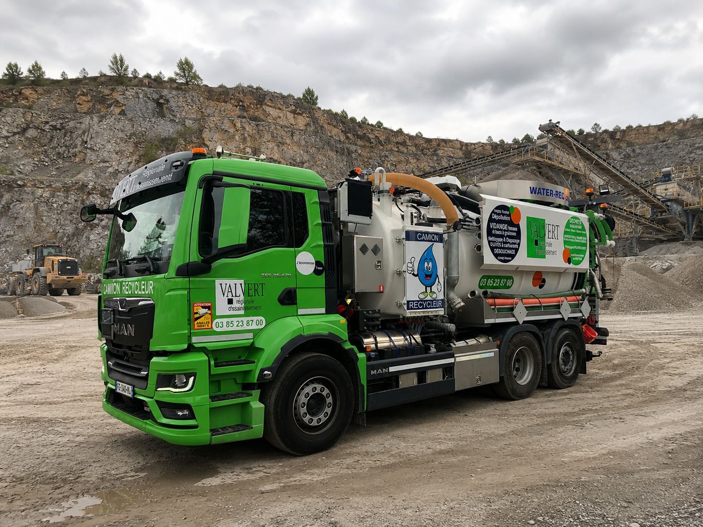 Un camion-citerne vert et blanc est stationné dans une rue étroite, devant un bâtiment, avec un tuyau bleu relié au sol.