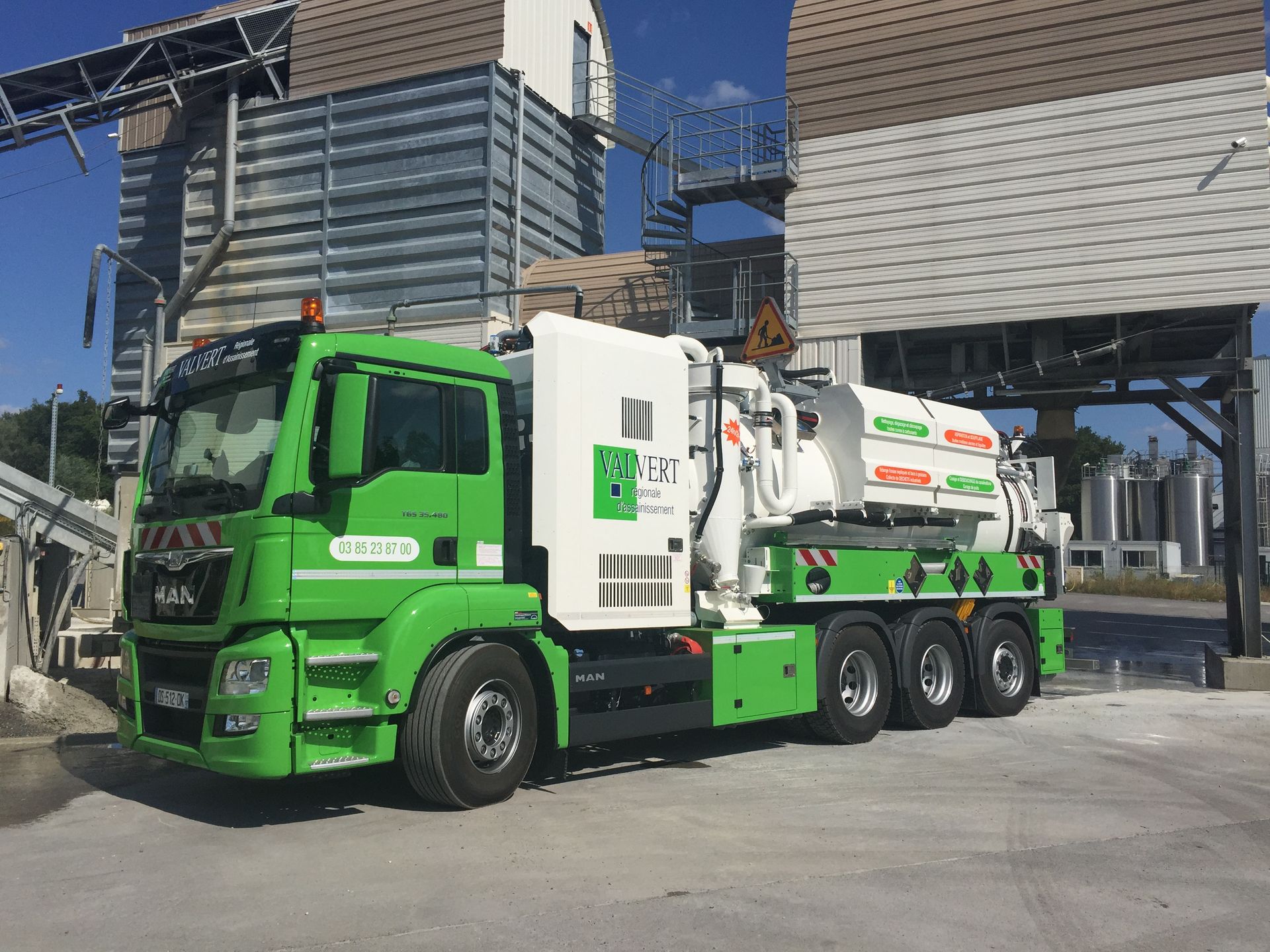 Camion industriel vert et blanc devant un bâtiment en béton, probablement une usine.