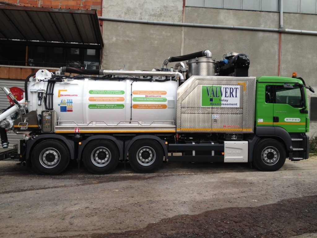 Camion aspirateur vert et blanc stationné devant un bâtiment industriel gris.