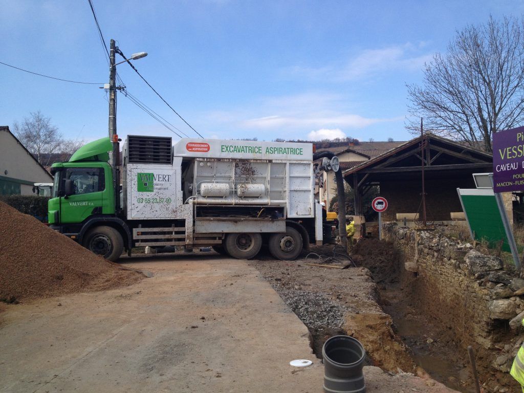 Camion vert et blanc, probablement un véhicule de travaux publics, stationné près d'un chantier sur une route étroite.