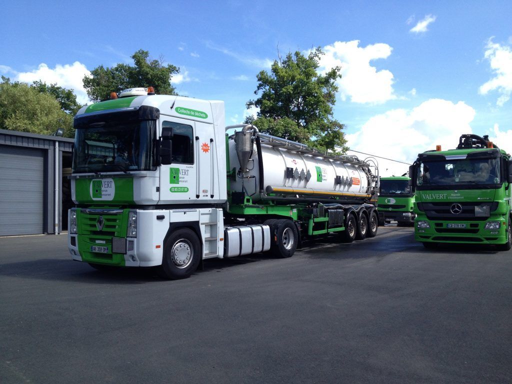 Un camion-citerne blanc et vert est stationné à côté d'autres camions sur une surface pavée, sur fond de ciel bleu.