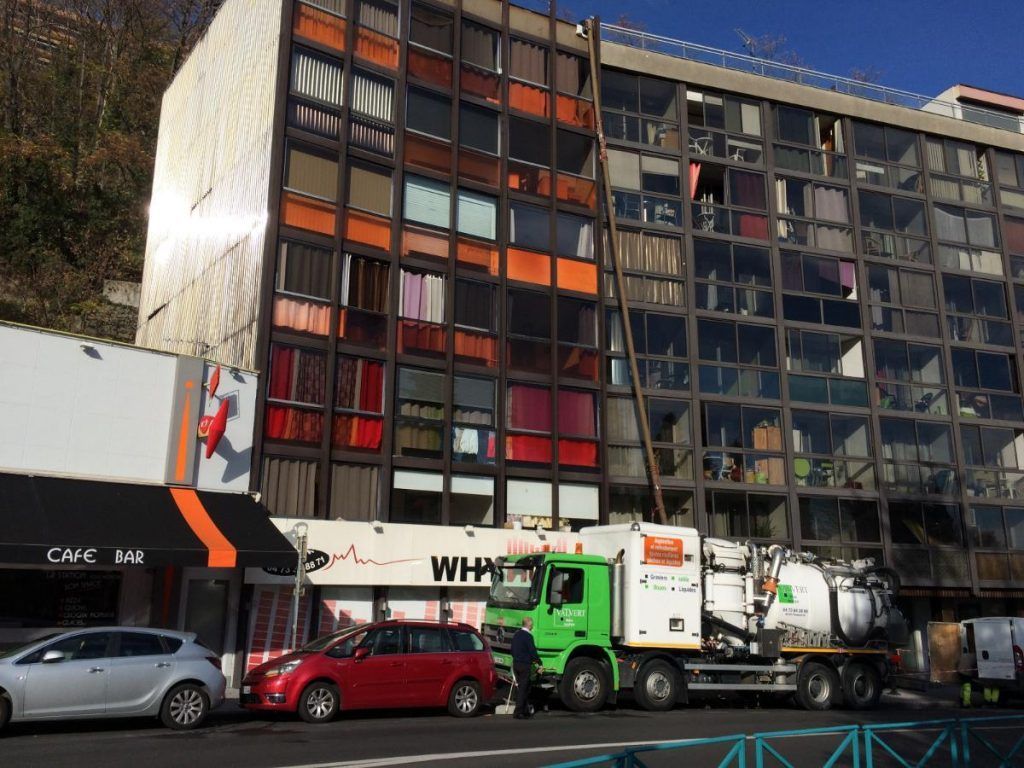 Immeuble avec de nombreuses fenêtres, véhicule utilitaire stationné dans la rue. On aperçoit également des voitures, un café et un arbre.