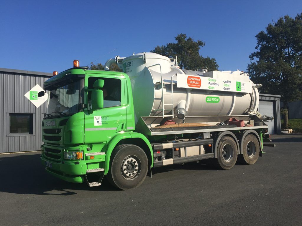 Un camion-citerne Scania vert avec une cuve blanche est stationné devant un bâtiment par une journée ensoleillée.