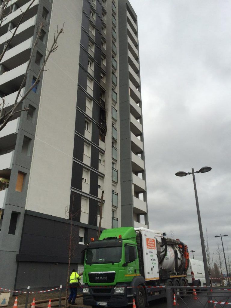 Immeuble de grande hauteur endommagé par un incendie, avec un camion vert garé en contrebas ; un pompier en gilet jaune est visible.