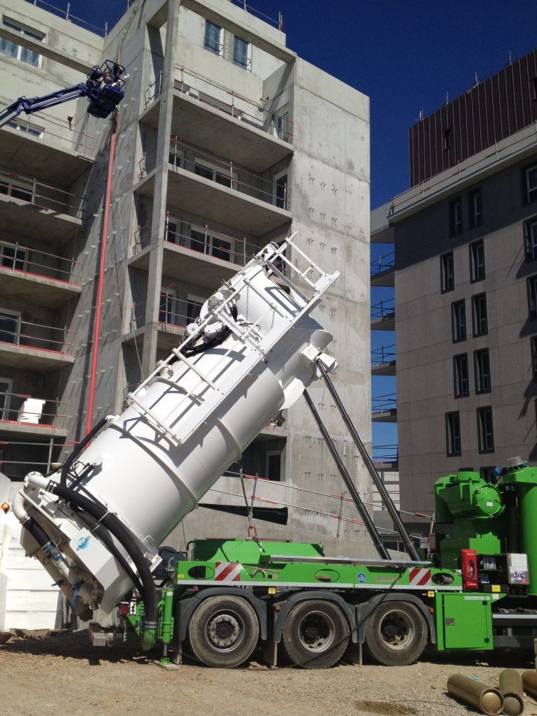 Camion aspirateur industriel blanc et vert incliné vers le haut sur un chantier. Bâtiments en béton en arrière-plan.