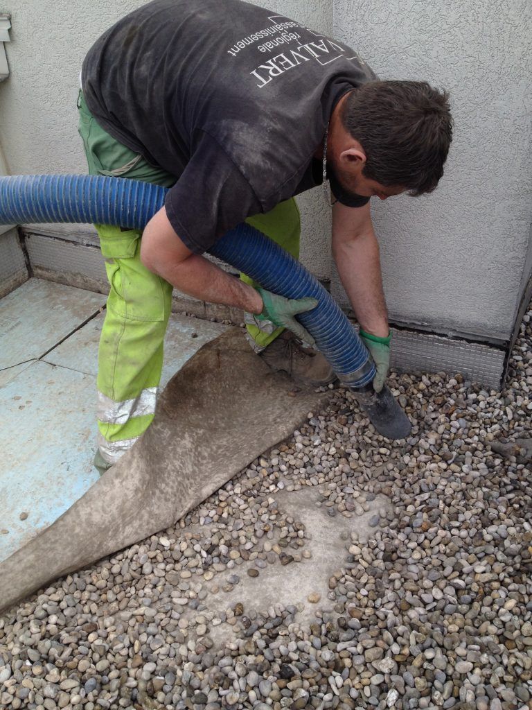Un homme en pantalon vert utilise un tuyau d'arrosage pour nettoyer le gravier sur un toit.