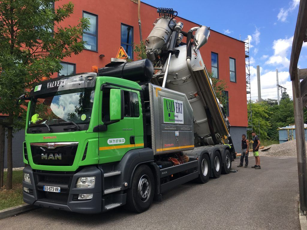 Un camion-citerne de ciment vert décharge devant un bâtiment rouge, deux ouvriers à proximité.