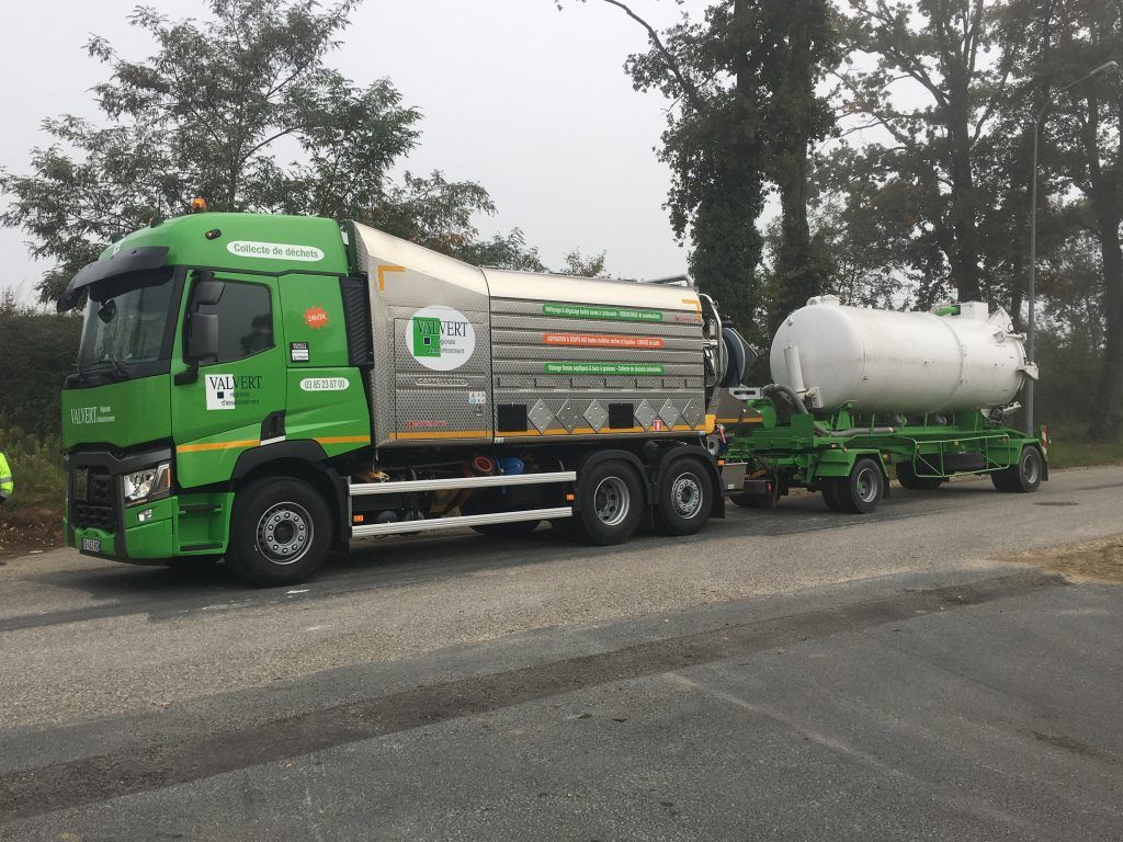 Camion vert tirant une semi-remorque citerne blanche sur une route asphaltée grise, arbres en arrière-plan.