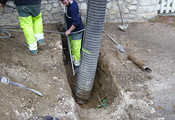 Deux ouvriers installent une grosse canalisation dans une tranchée. L'un d'eux tient un niveau et porte un équipement de sécurité vert.