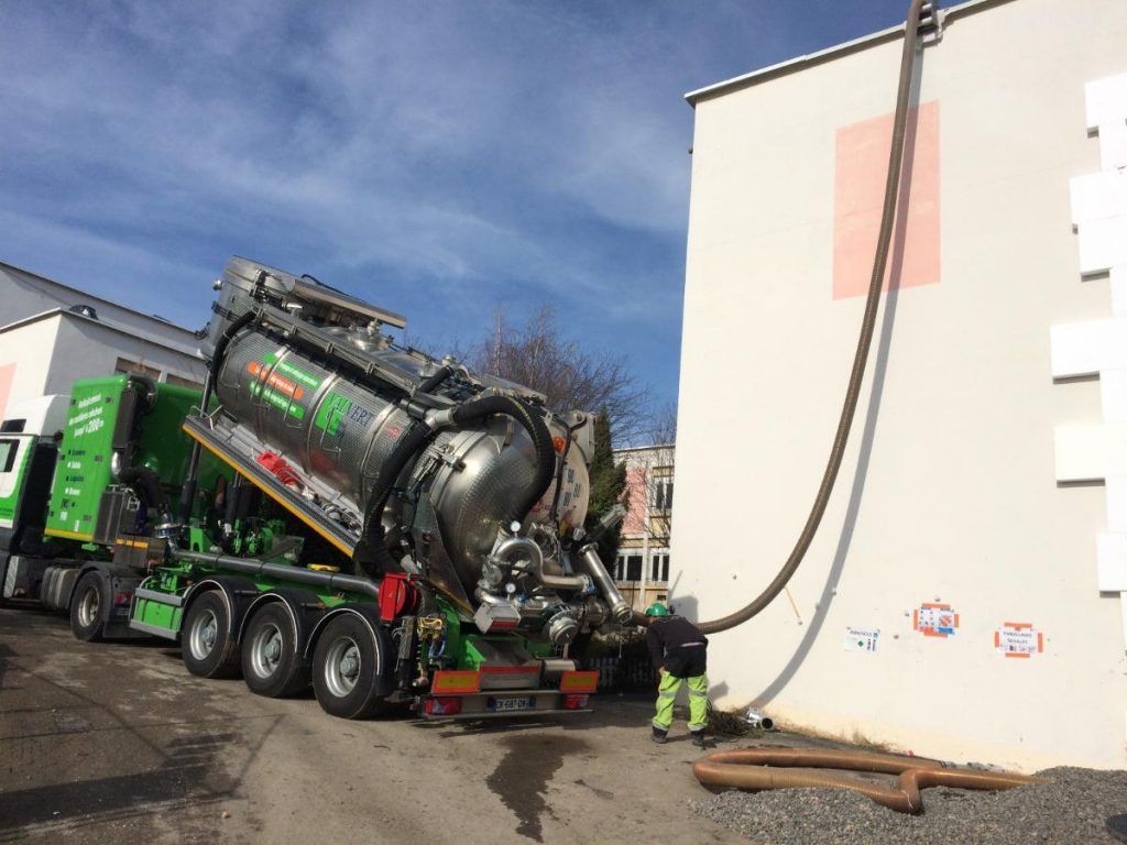 Un camion-citerne vert vide les déchets d'un bâtiment. Un employé en jaune manipule le tuyau.