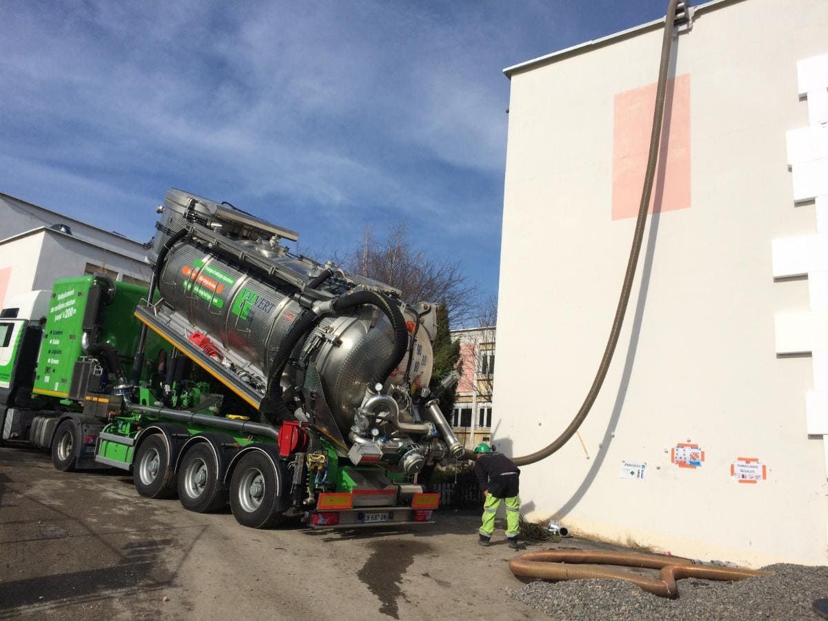 Un camion-citerne vert, sa citerne levée, vide un tuyau près d'un bâtiment.