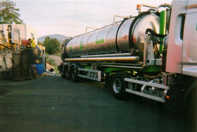 Camion-citerne stationné près d'une installation industrielle ; cuve métallique, éléments verts, tuyaux et canalisations.